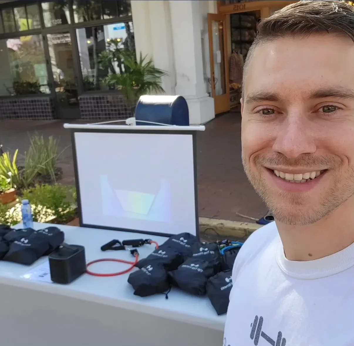 Laurence Larsen smiling in a white Tip Top Fitness T-shirt beside a display table with shirts and exercise equipment